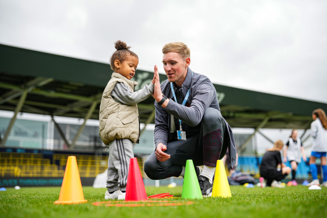 A coach kneels on a soccer field, giving a high-five to a child standing beside colorful cones. Both their faces are blurred. In the background, other children and adults are visible, likely participating in a sports event.