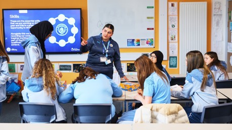 A woman leads a workshop with students wearing Manchester City clothing. Skills and the Manchester City logo are visible on the screen.