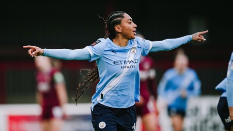 A soccer player in a Manchester City jersey celebrates with arms outstretched during a match.