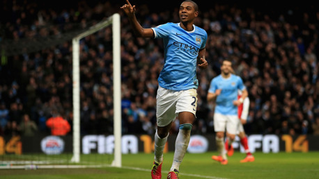A footballer from Manchester City celebrating after scoring a goal, wearing the team's blue jersey with the Etihad logo. The match is held at Etihad Stadium.