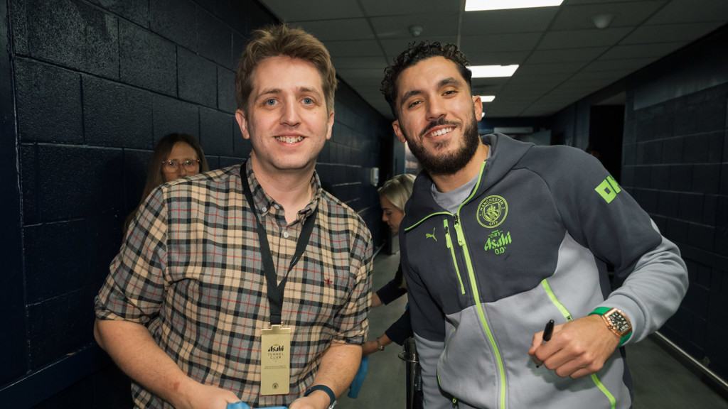 A guest wearing a plaid shirt with a VIP lanyard stands next to a Manchester City staff member in a team tracksuit in a hallway.