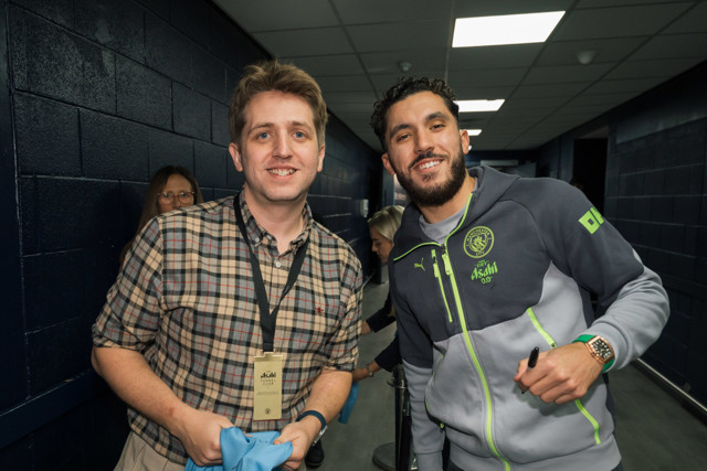 A guest wearing a plaid shirt with a VIP lanyard stands next to a Manchester City staff member in a team tracksuit in a hallway.