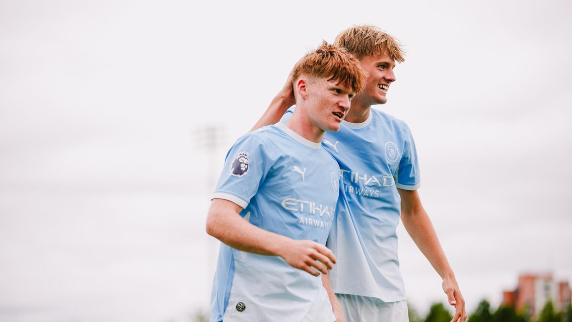 Two young male football players in light blue Manchester City kits celebrate on the field, with one player's arm around the other.