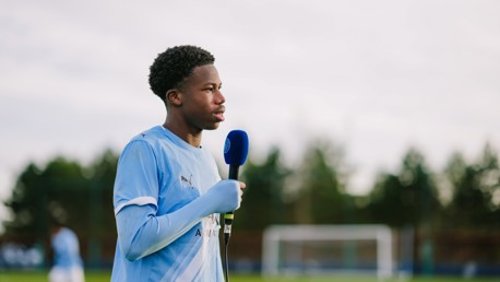 A Manchester City player holding a microphone wearing a light blue Puma jersey on a football field.
