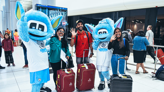 Two people posing with Manchester City's mascots Moonchester and Moonbeam in an airport terminal with luggage.