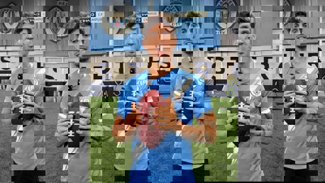 Three individuals holding American footballs wearing Manchester City jerseys in an indoor training facility with the Manchester City crest in the background.