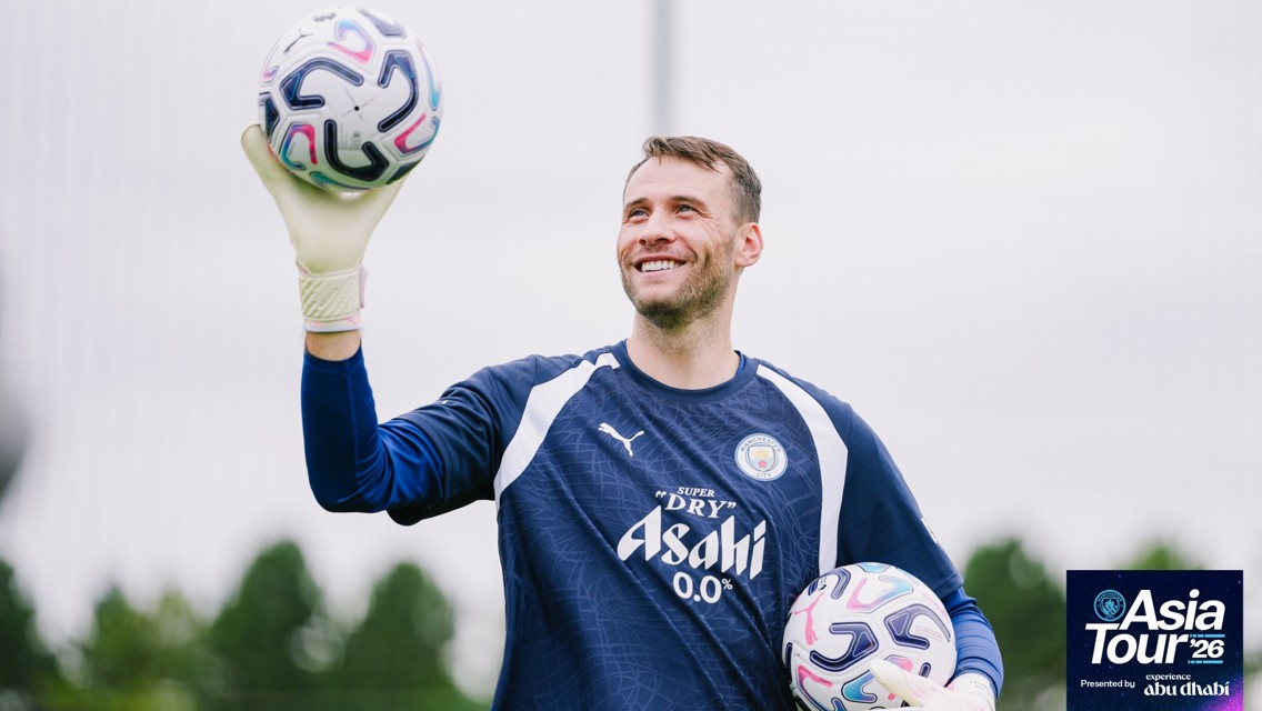 A goalkeeper in a Manchester City training kit smiling and holding two footballs during the Asia Tour 2026. The training is sponsored by Asahi and Abu Dhabi.
