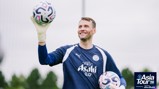 A goalkeeper in a Manchester City training kit smiling and holding two footballs during the Asia Tour 2026. The training is sponsored by Asahi and Abu Dhabi.
