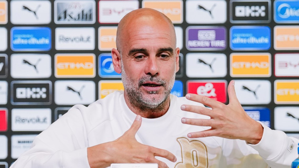 A manager at a press conference table with Manchester City logos in the background, wearing a white shirt and gesturing with hands.