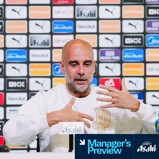 Un entrenador en una mesa de conferencia de prensa con los logotipos del Manchester City de fondo, vestido con una camiseta blanca y gesticulando con las manos.