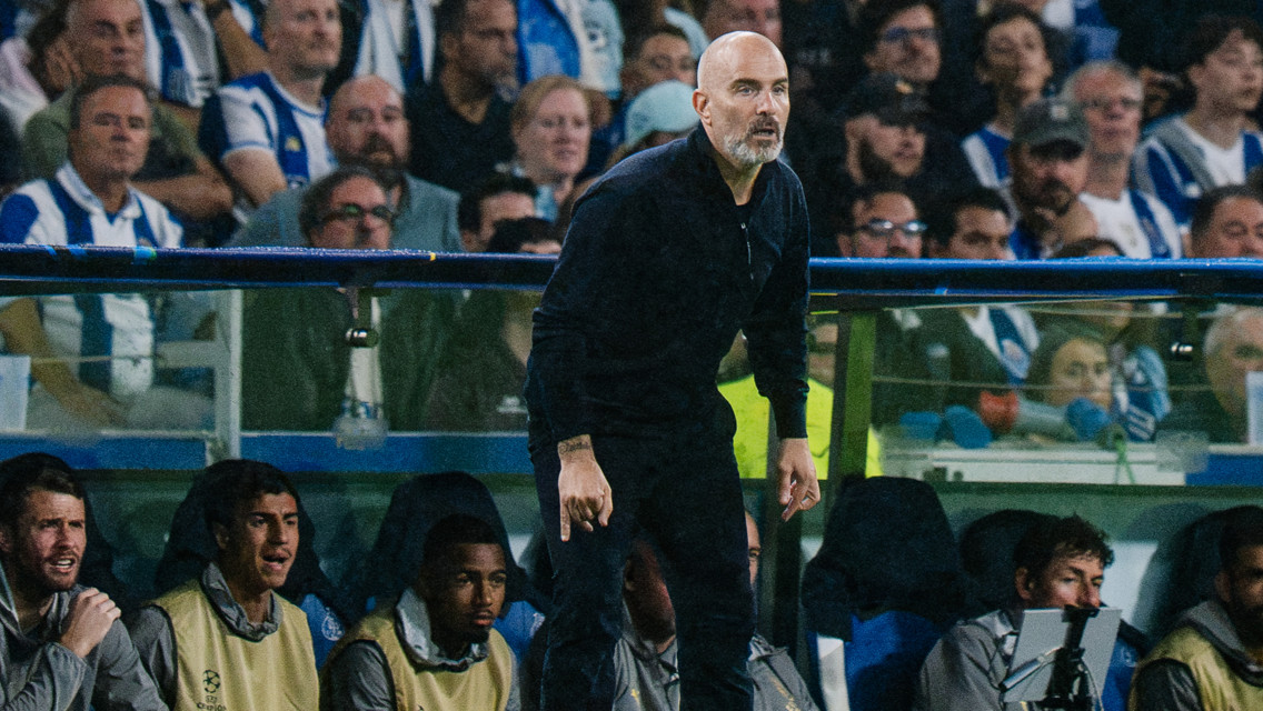 A football coach on the sideline during a match, with players seated behind him and a crowd in the background.