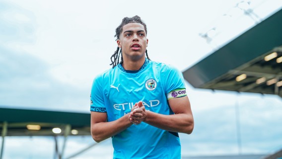 A person wearing a Manchester City football kit is clapping at a stadium. The sky is overcast.