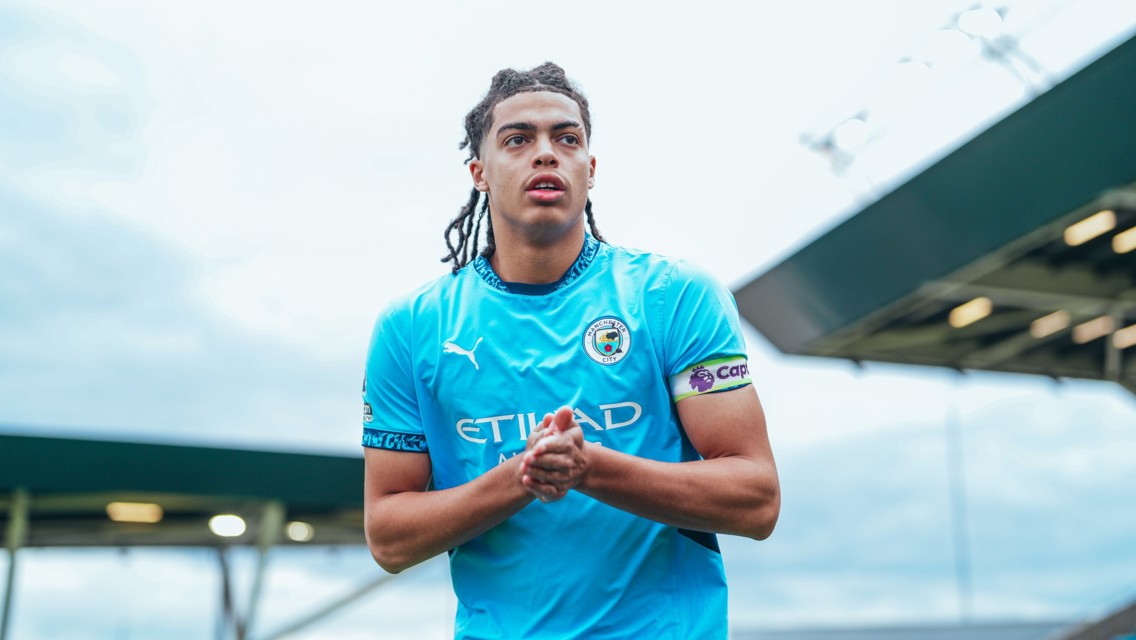 A person wearing a Manchester City football kit is clapping at a stadium. The sky is overcast.