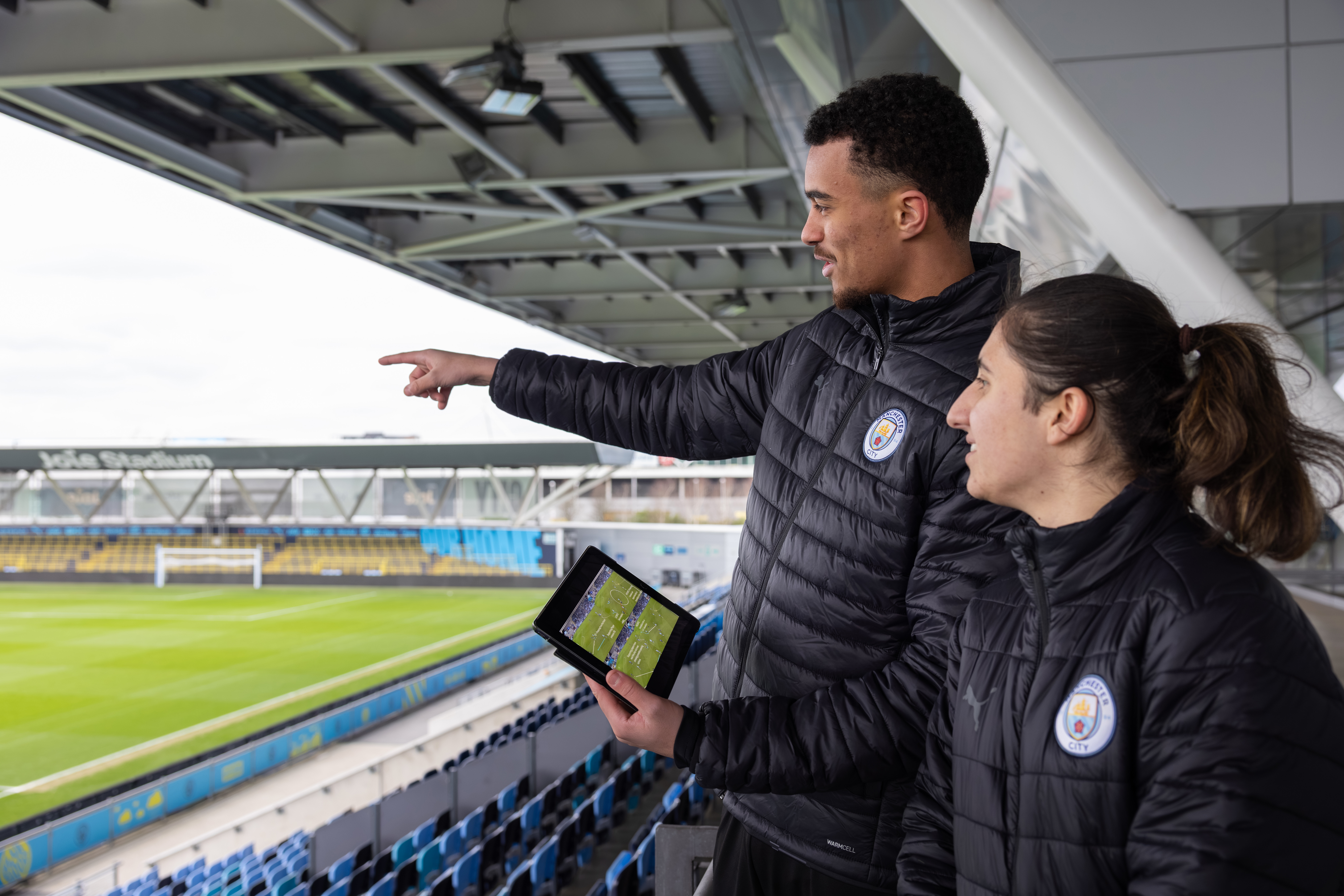 Two individuals in Manchester City jackets reviewing a football field with a tablet showing a tactical screen.