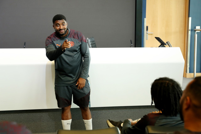 A Manchester City football player dressed in training gear is speaking and pointing towards an audience during a session, likely within a conference or training room environment.