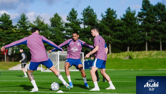 Three soccer players in purple Asahi Dry branded training jackets and blue shorts practice on a green training pitch with trees in the background.