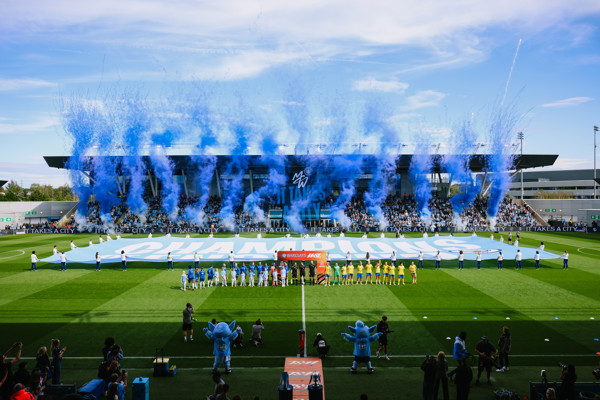 Players line up on a football field with a large blue banner and fans in the background. Blue smoke is launched into the air, celebrating a matchday event.