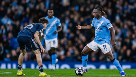 A Manchester City player in light blue kit dribbling a ball while an opponent in blue kit prepares to defend during a soccer match.