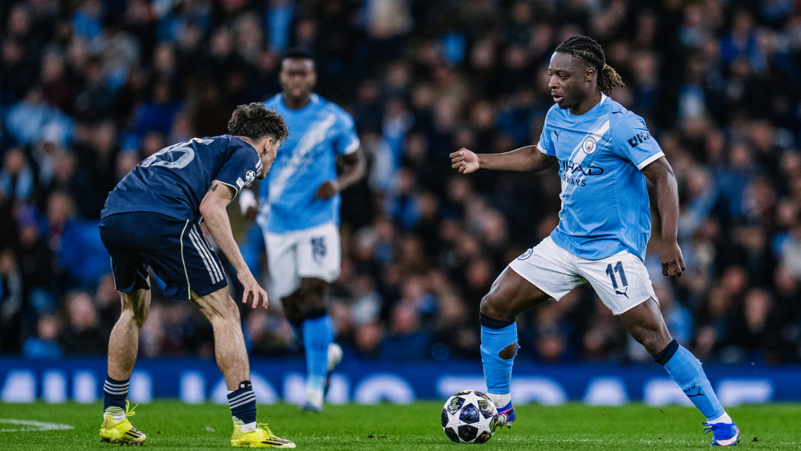 A Manchester City player in light blue kit dribbling a ball while an opponent in blue kit prepares to defend during a soccer match.