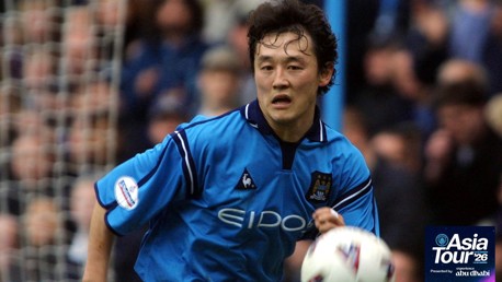 A football player in a Manchester City jersey during a game action shot, wearing a light blue kit and running with the ball. The Asia Tour 2026 logo is visible in the corner.