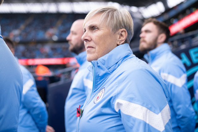A Manchester City staff member wearing a blue jacket with the club's logo stands at the Etihad Stadium.