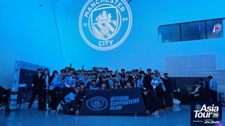 A group photo of Manchester City Official Supporters Club holding a banner at the Asia Tour '26 event. The Manchester City crest is projected on the wall behind them.