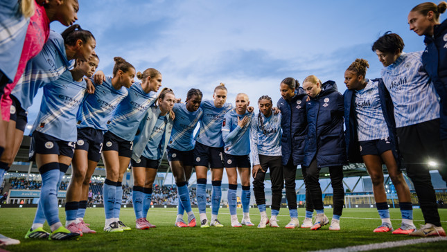 Manchester City women's football team in a pre-match huddle wearing blue Etihad Airways jerseys on a football field.