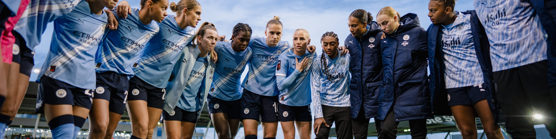 Manchester City women's football team in a pre-match huddle wearing blue Etihad Airways jerseys on a football field.