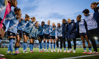 Manchester City women's football team in a pre-match huddle wearing blue Etihad Airways jerseys on a football field.