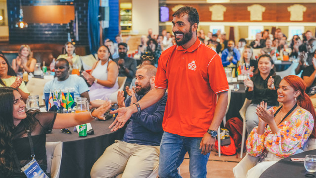 A man in a red polo shirt with a logo saying Oscar Foundation shakes hands with a woman in black at a seated event with diverse attendees and small international flags on tables.