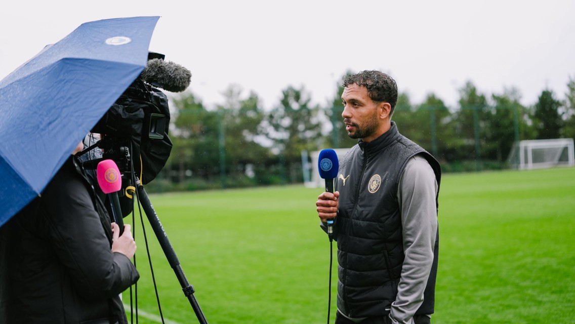 A man in a black vest is being interviewed on a football field holding a blue microphone. A camera person with a pink microphone under a blue umbrella records the interview.