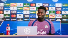 A Manchester City player at a press conference with logos of sponsors like FedEx and Walkers, and UEFA Champions League branding in the background.