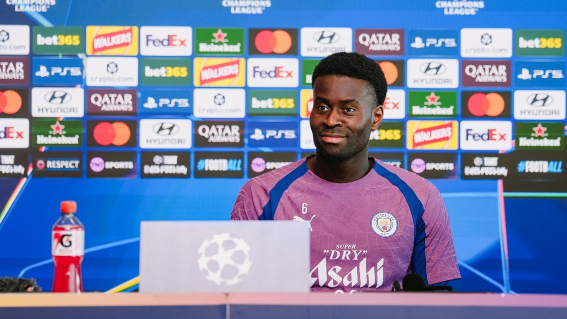 A Manchester City player at a press conference with logos of sponsors like FedEx and Walkers, and UEFA Champions League branding in the background.