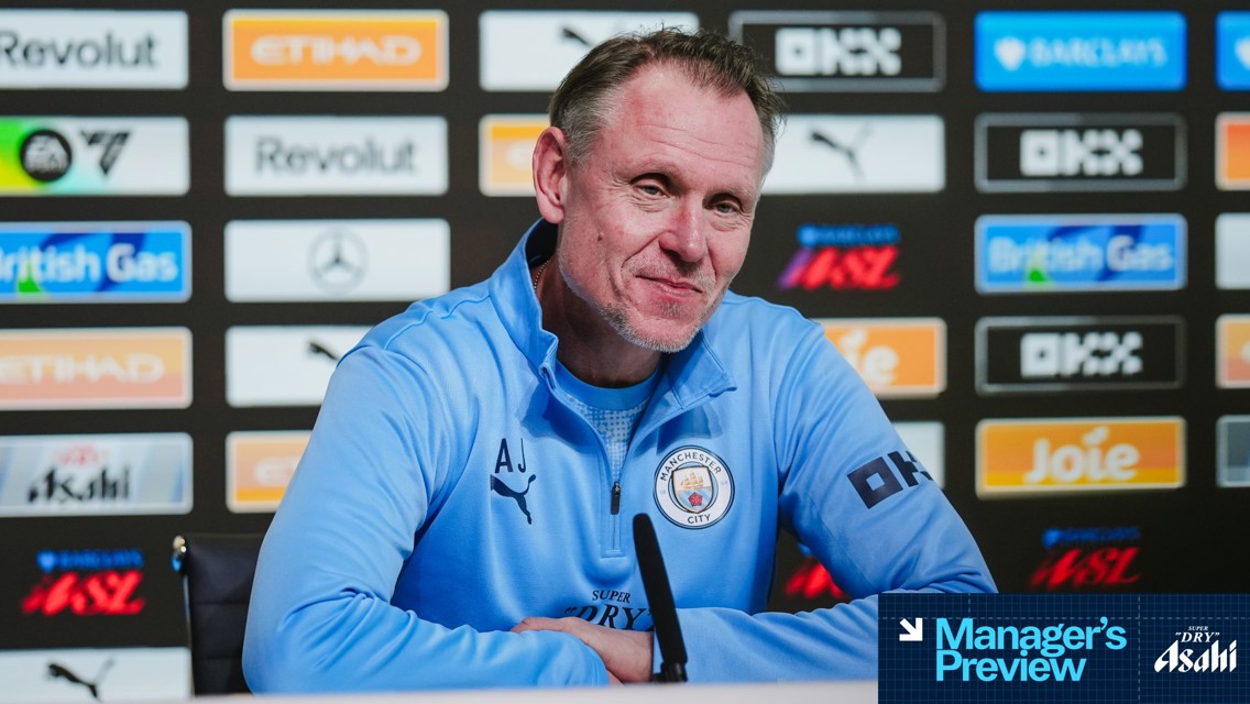 A person wearing a Manchester City tracksuit with the initials 'AJ' sits at a press conference table with logos in the background.