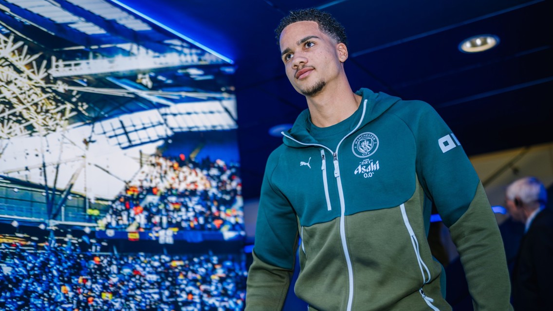 A Manchester City player wearing a team tracksuit enters the tunnel at Etihad Stadium. Blurred background includes stadium seating and lighting.