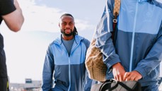 A person walking in a blue Manchester City tracksuit, holding luggage, with a second person partly visible carrying a backpack and bag.