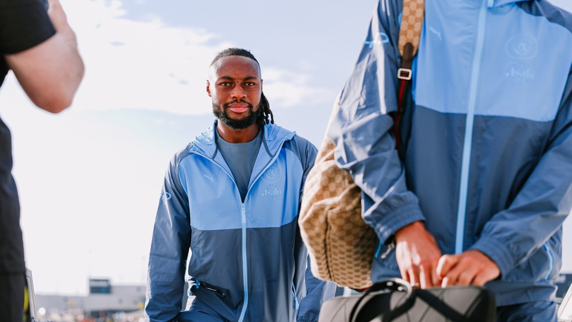 A person walking in a blue Manchester City tracksuit, holding luggage, with a second person partly visible carrying a backpack and bag.