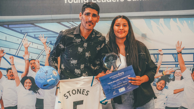 Two people hold a signed Manchester City football, a John Stones jersey, an award, and a plaque at the Cityzens Giving Young Leaders Summit event.