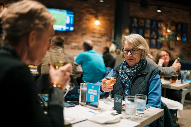 People enjoying drinks at a restaurant with a blurred view of a televised sports game in the background.