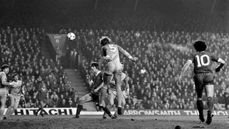 Black and white image of a football match, showing a player jumping to head the ball in front of a crowded stadium. Players from two teams, one with number 5 and another with number 10, are visible.