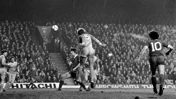 Black and white image of a football match, showing a player jumping to head the ball in front of a crowded stadium. Players from two teams, one with number 5 and another with number 10, are visible.