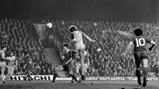 Black and white image of a football match, showing a player jumping to head the ball in front of a crowded stadium. Players from two teams, one with number 5 and another with number 10, are visible.