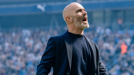 A bald man with a beard wearing a black jacket and blue shirt is yelling with his mouth open, standing in a stadium with a blurry crowd in the background.