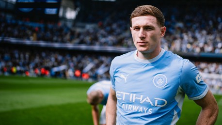 A football player in a Manchester City uniform standing on the field at Etihad Stadium. The player is wearing a light blue jersey with the Etihad Airways logo and Puma branding, while a blurred crowd fills the stadium stands.