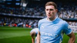A football player in a Manchester City uniform standing on the field at Etihad Stadium. The player is wearing a light blue jersey with the Etihad Airways logo and Puma branding, while a blurred crowd fills the stadium stands.