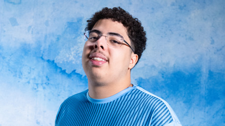 A young man with glasses wearing a blue shirt stands in front of a blue textured background.