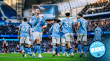 Players from Manchester City celebrate a goal at a football match at Etihad Stadium. Visible players have their names on the jerseys, and fans are in the background.