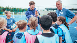 A group of young soccer players, coached by staff in Manchester City gear, are gathered on a soccer field, receiving instruction.