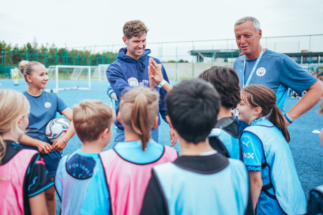 A group of young soccer players, coached by staff in Manchester City gear, are gathered on a soccer field, receiving instruction.