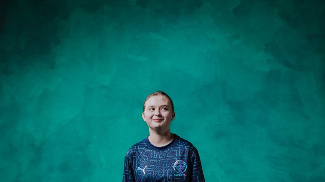 A person wearing a Manchester City Esports shirt standing against a green textured background. The face is blurred.